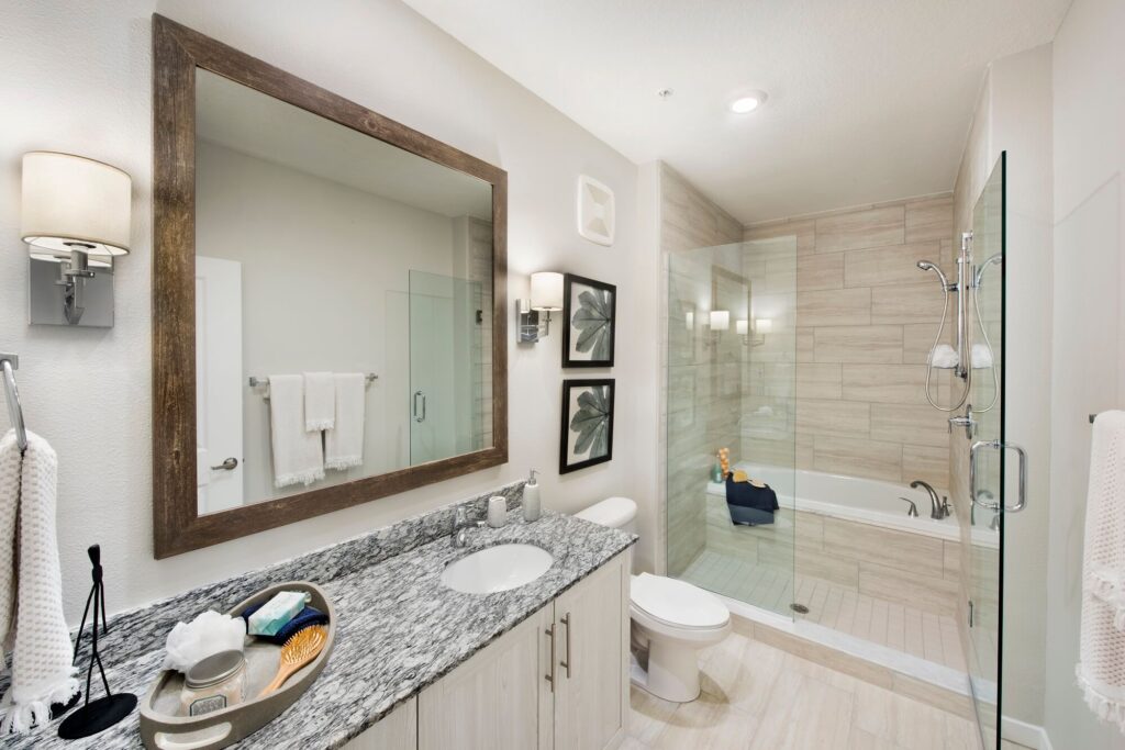 Modern bathroom with a large mirror above a granite countertop, sink, and light fixtures. There are towels, a toilet, framed artwork, and a spacious shower next to a bathtub with beige tiles.