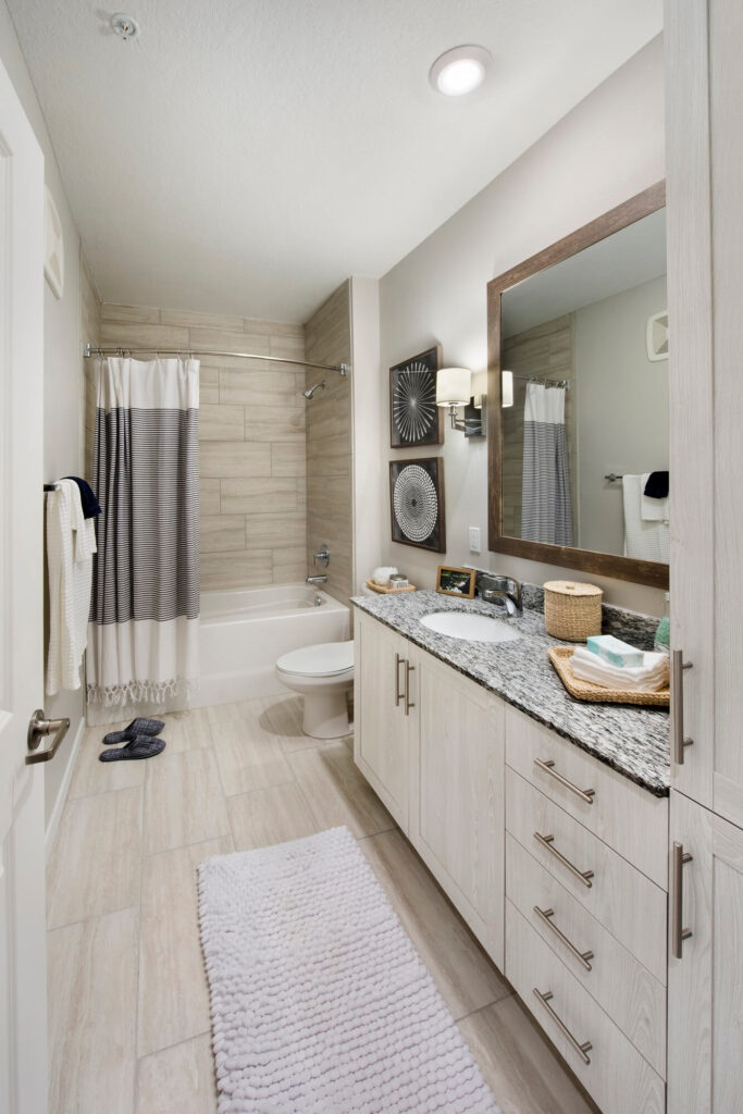 Modern bathroom with light wood cabinets, granite countertop, large mirror, white tub with gray and white shower curtain, tiled walls and floor, wall decor, towels, and a textured white rug on the floor.