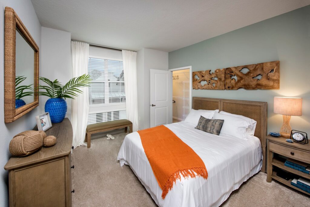 A modern bedroom with a bed covered in white linens and an orange throw, wooden furniture, a decorative wooden wall art, a blue lamp, potted plant, window with white curtains, and beige carpet.