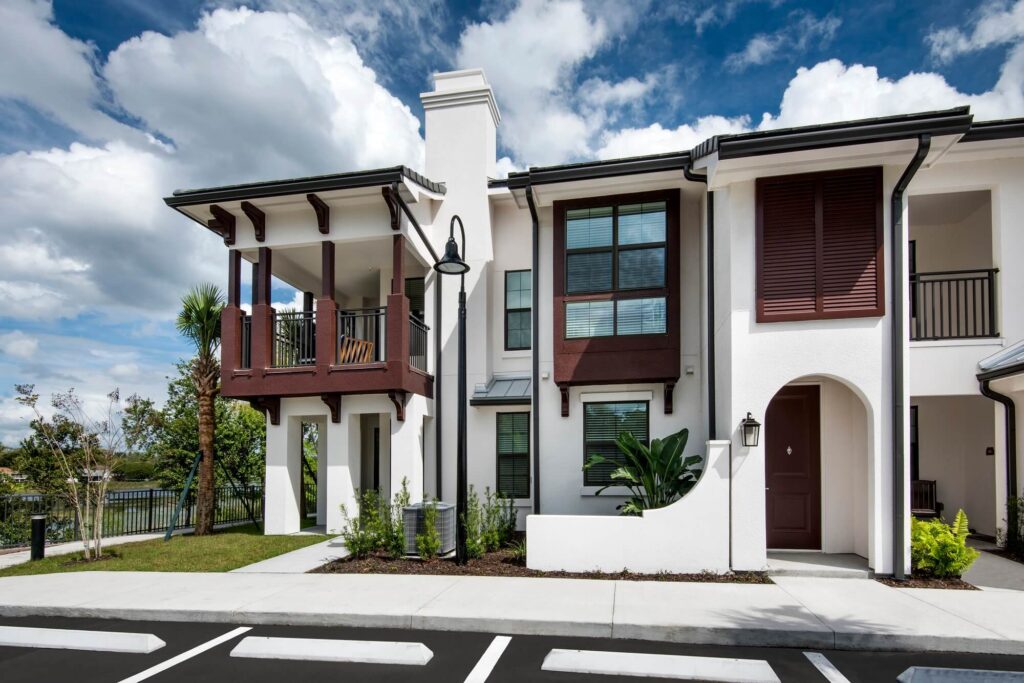 Modern townhouse with white exterior, dark brown accents, large windows, a covered balcony, and landscaped greenery beside a parking lot under a partly cloudy sky.