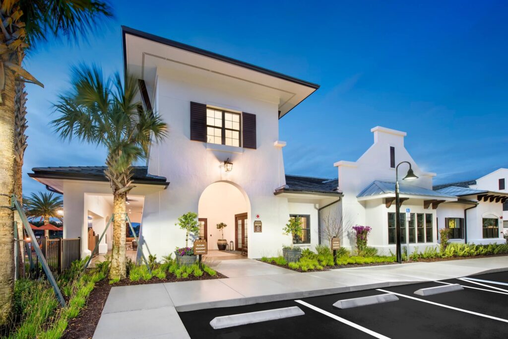 A modern, white stucco building with a tall archway entrance, palm trees, landscaped greenery, and an empty parking lot in front, shown at dusk with a clear blue sky.