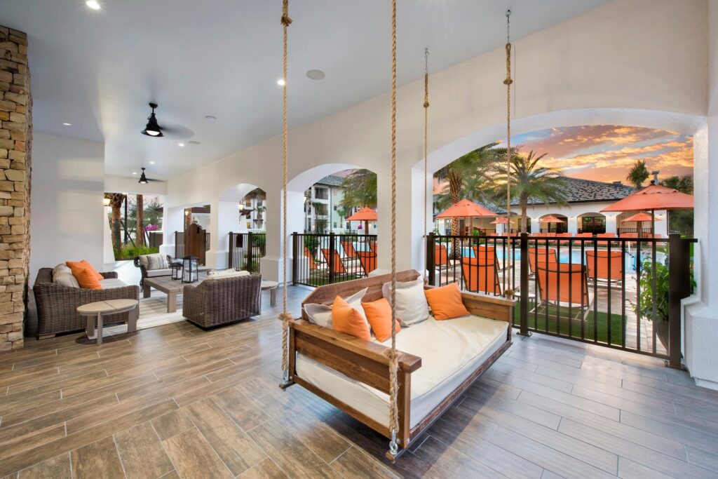 Covered patio with a wooden porch swing, wicker seating with orange cushions, and view of a pool area with umbrellas and palm trees at sunset. The space has wood-look tile flooring and stone pillars.