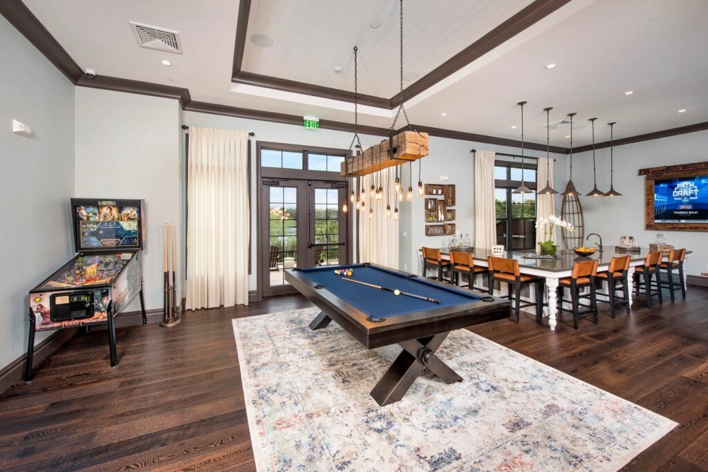 A spacious game room with a pool table on a decorative rug, a pinball machine, and a long dining table with chairs under modern pendant lights, featuring large windows and wood floors.
