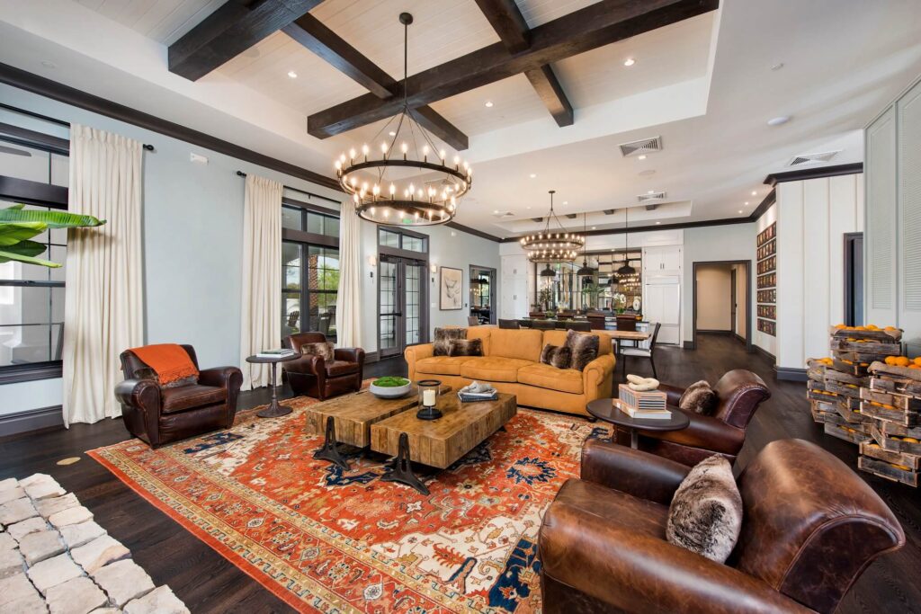 Spacious living room with exposed wood beams, two large chandeliers, tan sofa, leather armchairs, a red-patterned rug, coffee tables, and large windows with cream curtains. The space feels bright and elegant.