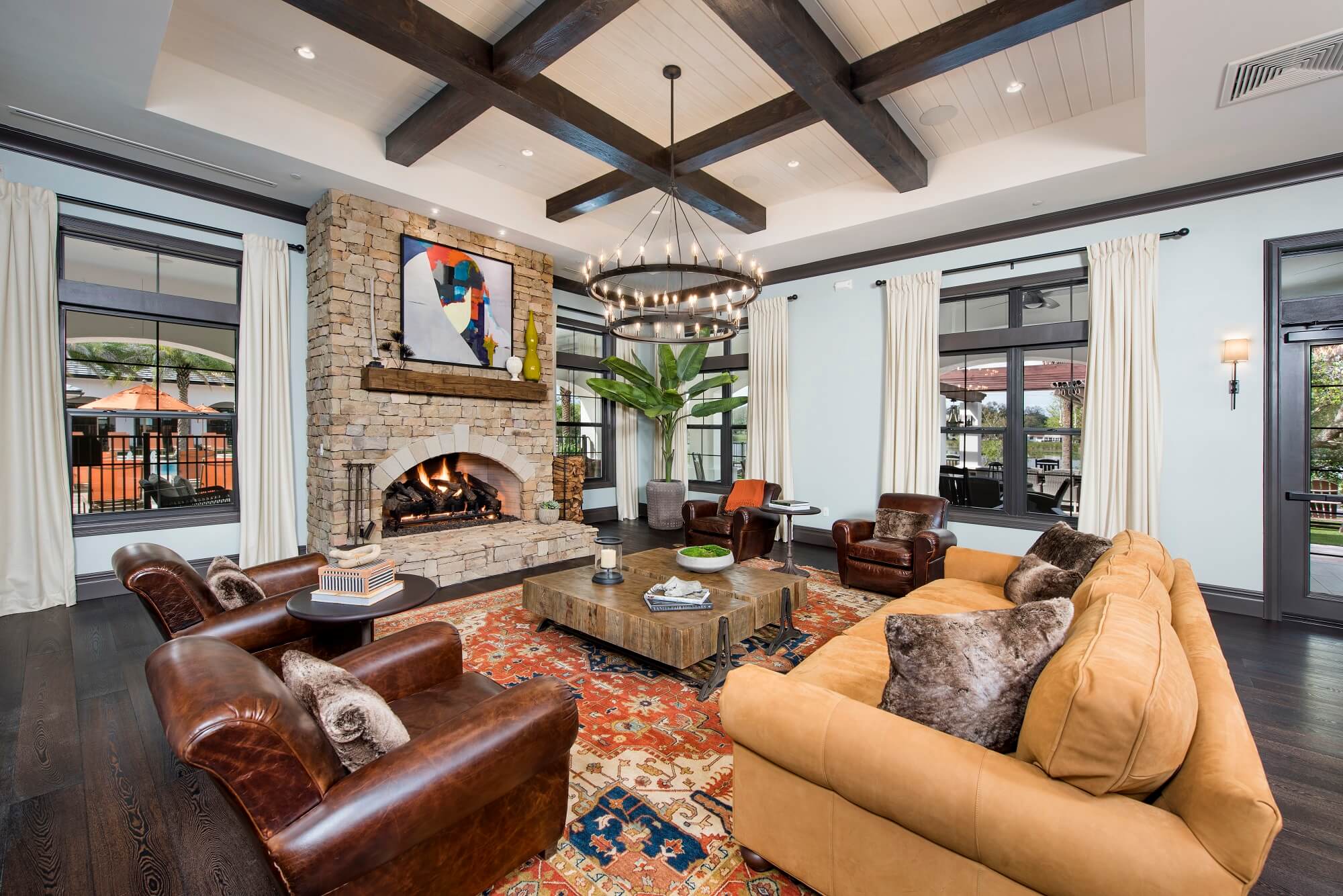 A spacious living room with a stone fireplace, modern art above it, leather armchairs, a tan sofa, a wooden coffee table, an ornate rug, a large plant, and big windows with white curtains letting in natural light.