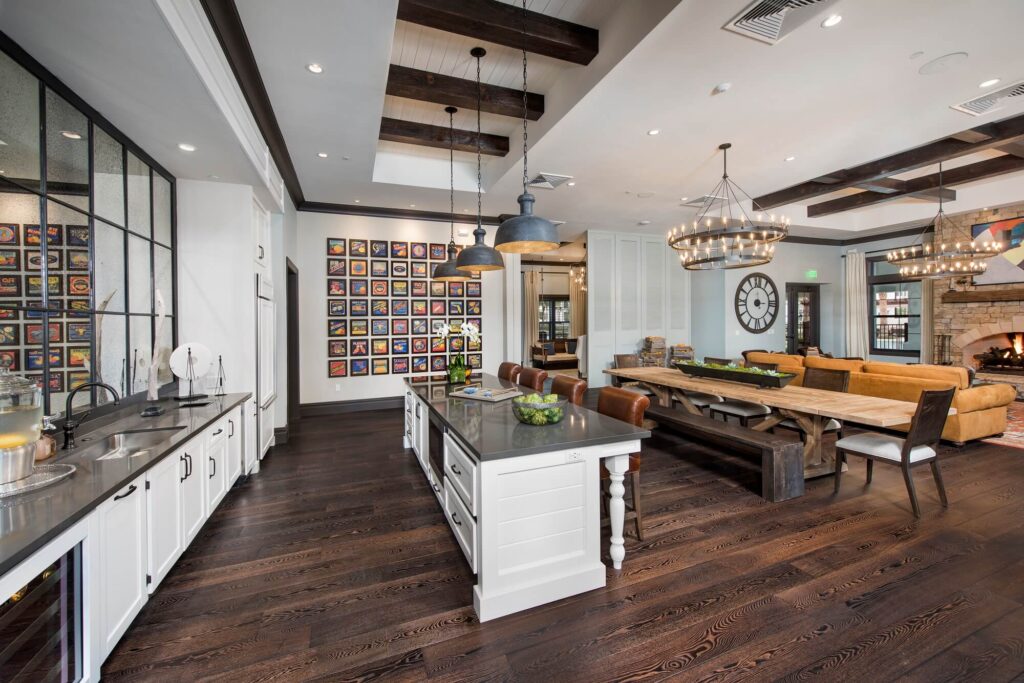 Spacious modern kitchen and living area with dark wood floors, white cabinetry, a large island with barstools, farmhouse tables, chandeliers, a wall of colorful framed art, and a stone fireplace in the background.