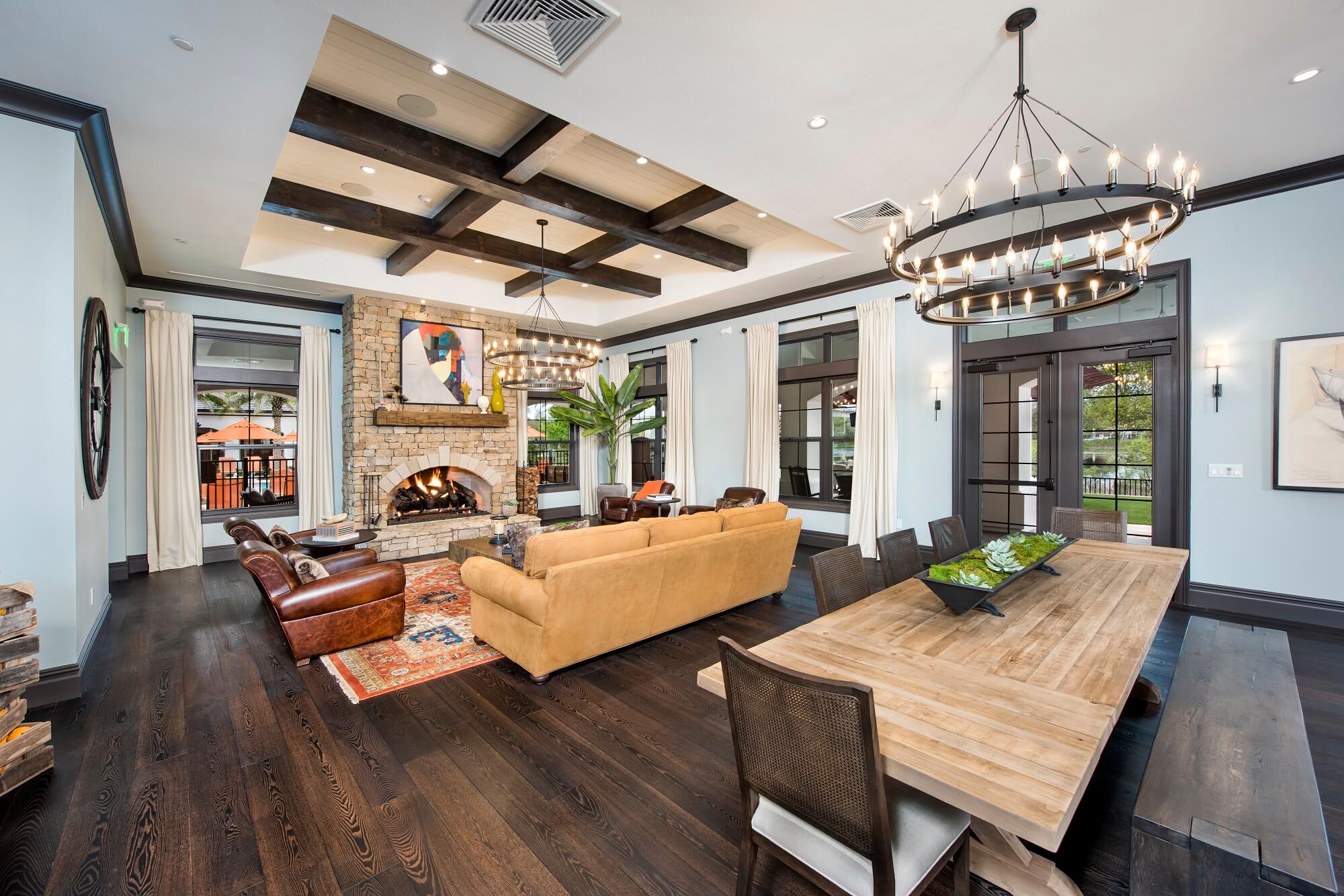 Spacious living and dining area with dark wood floors, exposed ceiling beams, a stone fireplace, large windows with white curtains, two modern chandeliers, and a mix of brown leather and tan sofas.