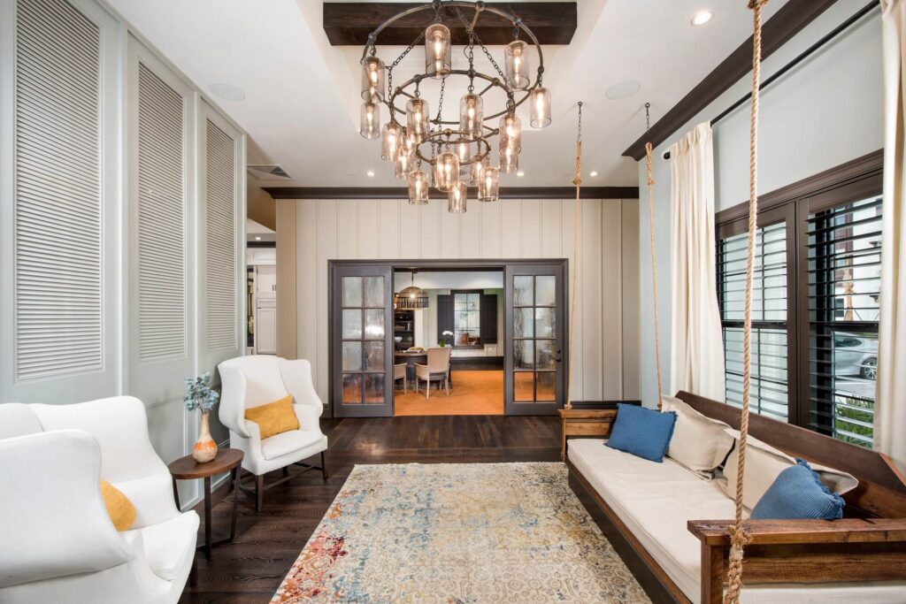 A stylish living room with two white armchairs, a rustic wooden swing with cushions, a patterned rug, a modern chandelier with exposed bulbs, and large windows with black shutters. Double doors open to another room.