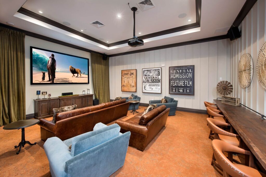 A stylish home theater room with leather and blue velvet seating facing a large screen, film posters on the walls, a projector on the ceiling, and tables along the side wall.