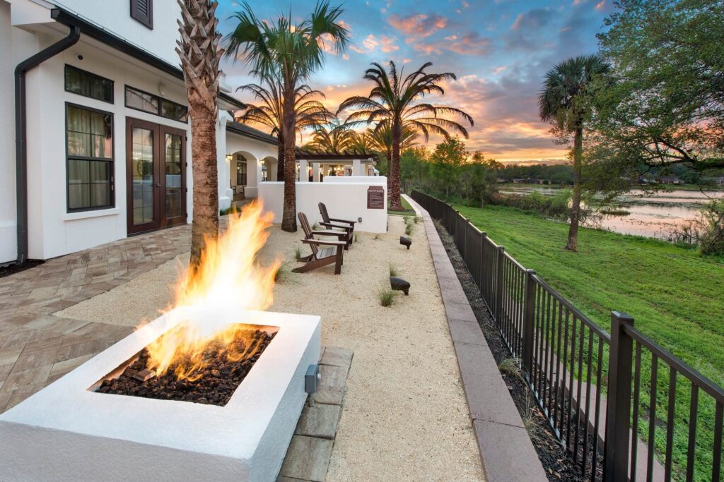 A modern patio with a fire pit, Adirondack chairs, palm trees, and a view of a grassy landscape and river at sunset. The house features large windows and white walls.
