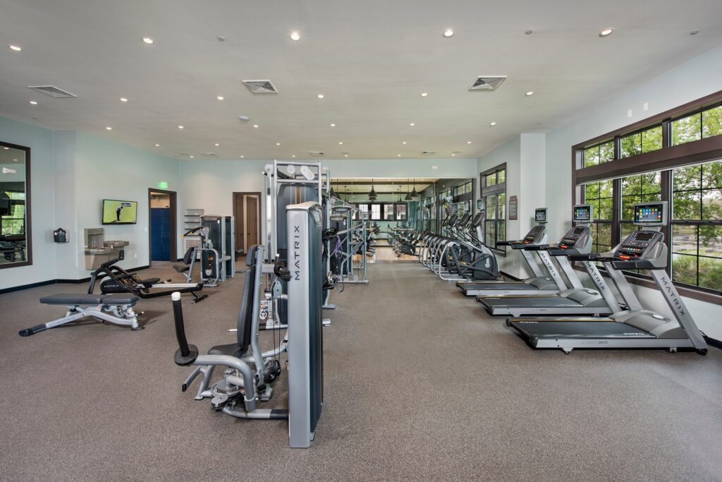 Modern gym with various exercise equipment, including treadmills, stationary bikes, weight machines, and benches. Large windows let in natural light, and mirrors line the back wall. The space is clean and organized.