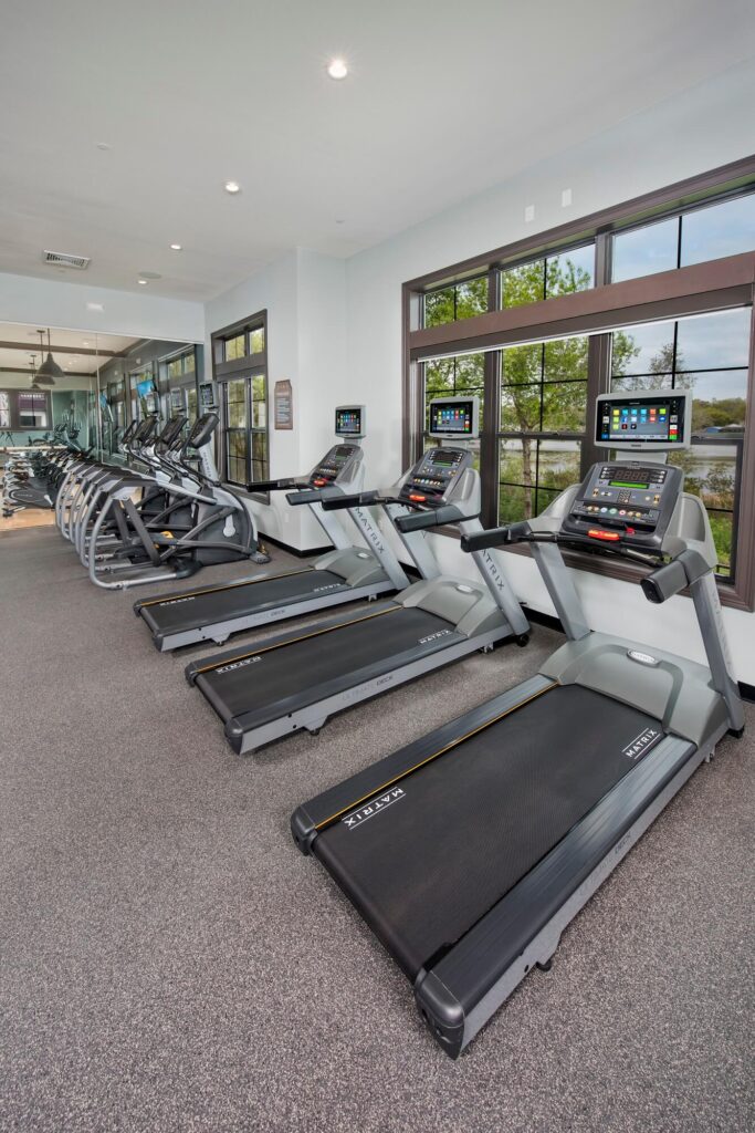 A modern gym with several treadmills and elliptical machines lined up next to large windows, allowing natural light to fill the spacious room.