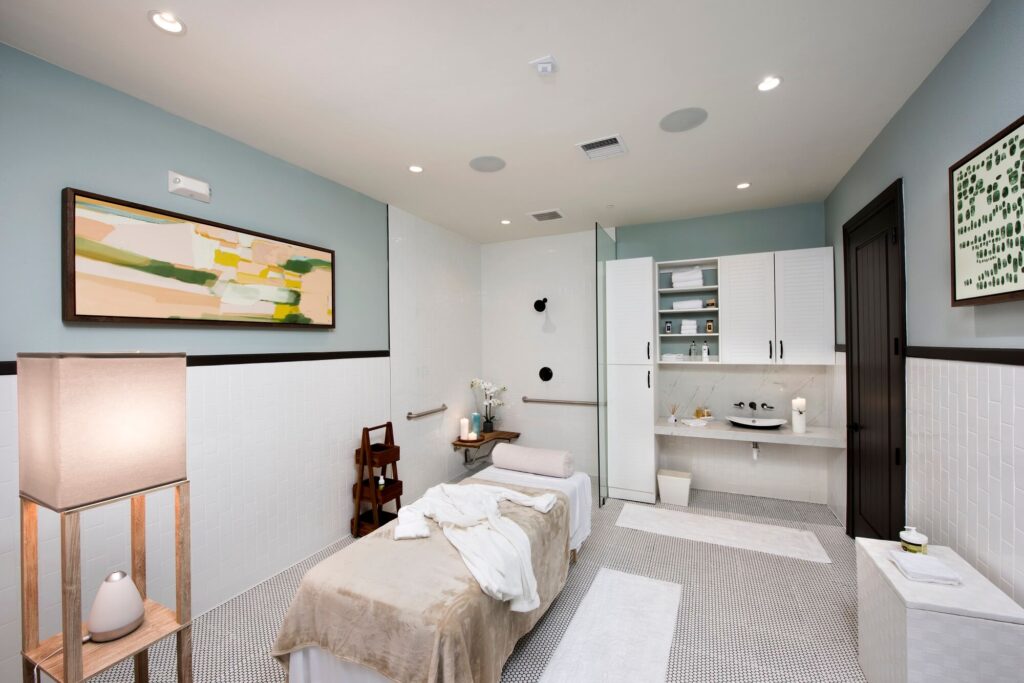 A modern spa treatment room with a massage table, towels, a floor lamp, shelves with spa products, and contemporary artwork on light blue and white walls. The space is clean and calming.