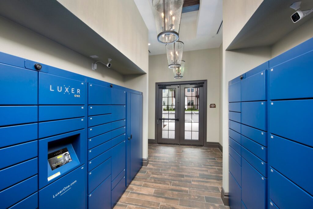 A modern package locker room with blue Luxer One lockers lining both sides, a touchscreen interface on the left, wood-look tile flooring, and glass double doors leading outside. Three pendant lights hang from the ceiling.