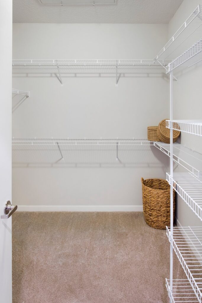 A clean, empty walk-in closet with beige carpet, white wire shelves along the walls, and two woven baskets placed on the floor and a shelf. The closet has white walls and a white door partially open.