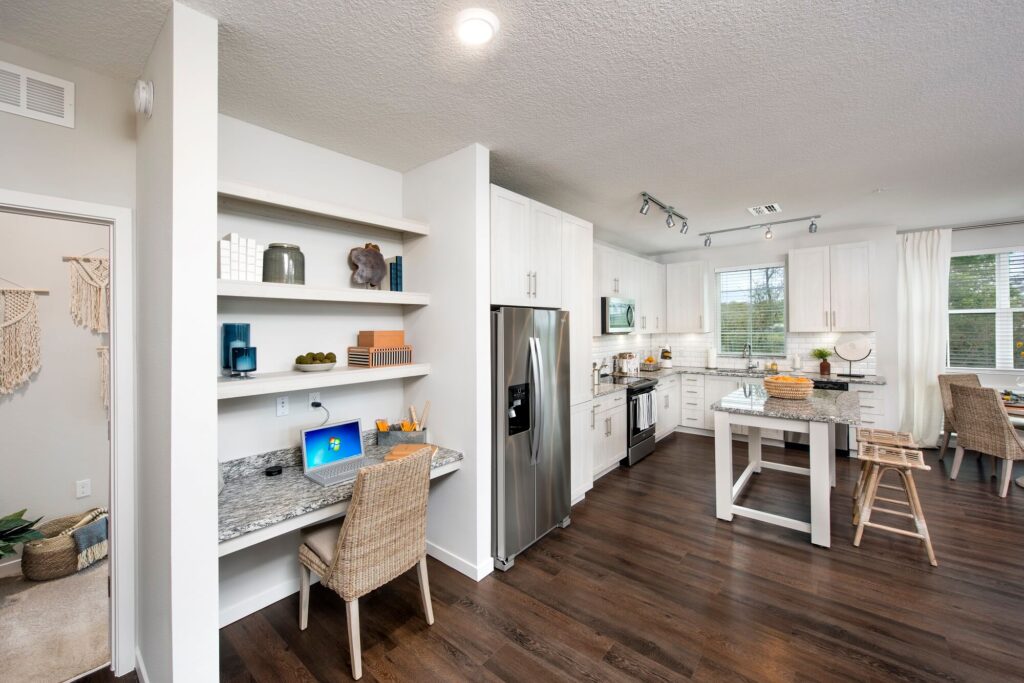Modern apartment with an open kitchen, stainless steel appliances, island with barstools, light cabinetry, and a small built-in desk workspace with a laptop and shelves, all featuring wood flooring and bright natural light.