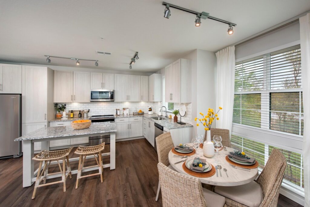 Modern kitchen with white cabinets, granite countertops, stainless steel appliances, and a small dining table set for four by large windows with white curtains. Two woven stools sit at the kitchen island.