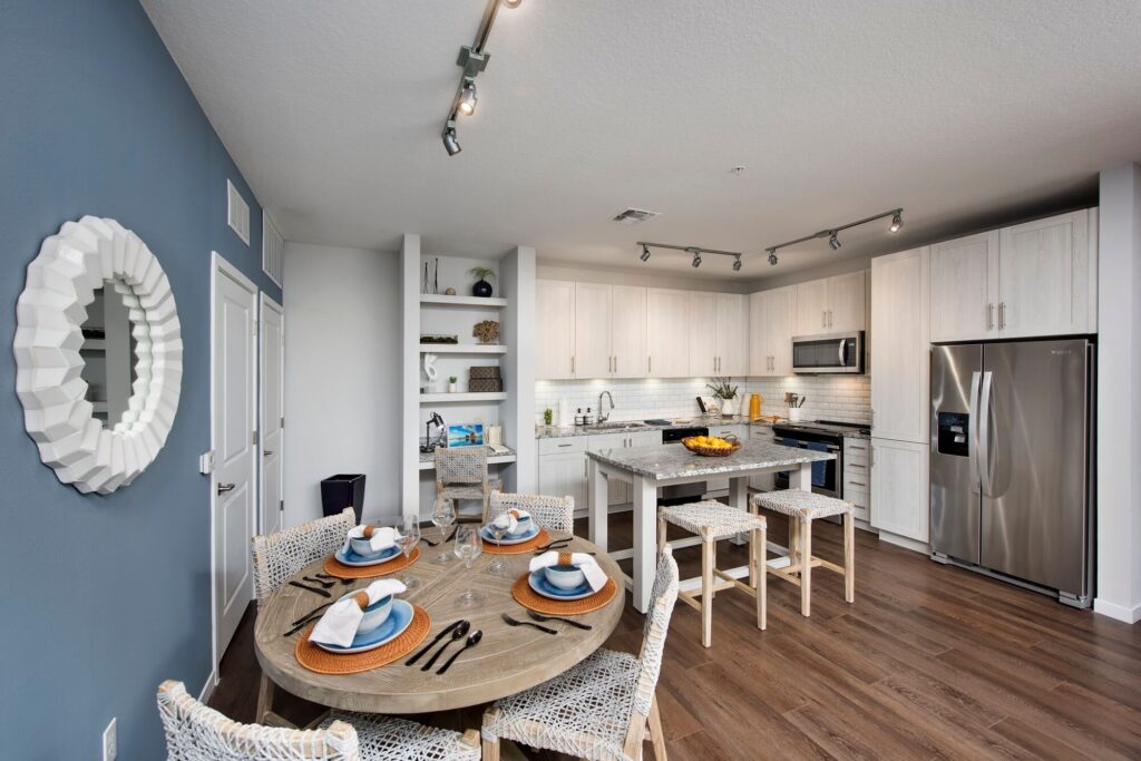 Modern kitchen and dining area with a round wooden table set for four, white cabinetry, stainless steel appliances, wooden stools at a kitchen island, and wood flooring. A blue accent wall with a round mirror is on the left.