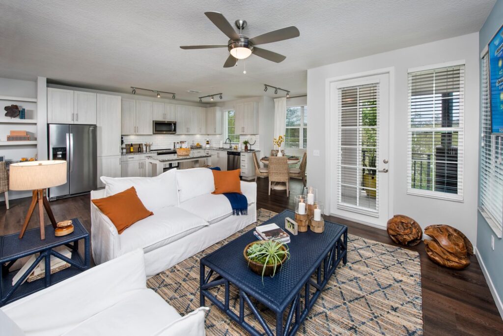 Modern open-concept living room and kitchen with white cabinetry, stainless steel appliances, white sofas with orange and blue pillows, blue tables, a patterned rug, and large windows letting in natural light.