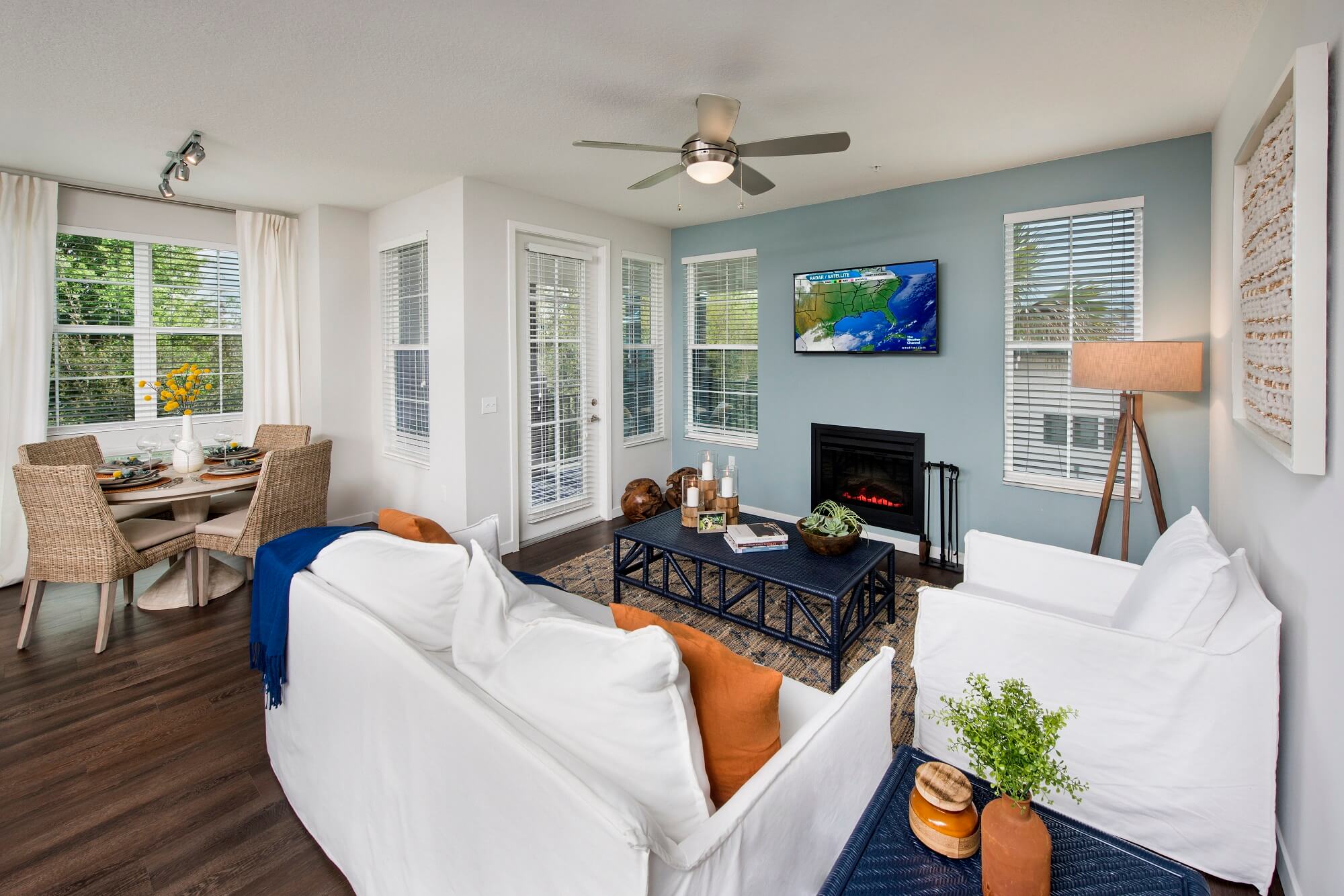 Modern living room with white sofas, orange pillows, a coffee table, and a fireplace under a wall-mounted TV. Dining area with wicker chairs is visible, and large windows let in natural light.