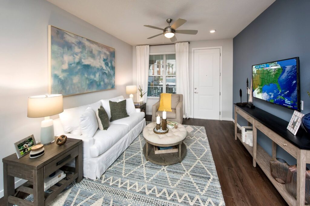 A modern living room with a white sofa, patterned rug, round coffee table, armchair, large wall art, TV displaying a weather map, and wooden furniture, all under soft lighting and a ceiling fan.