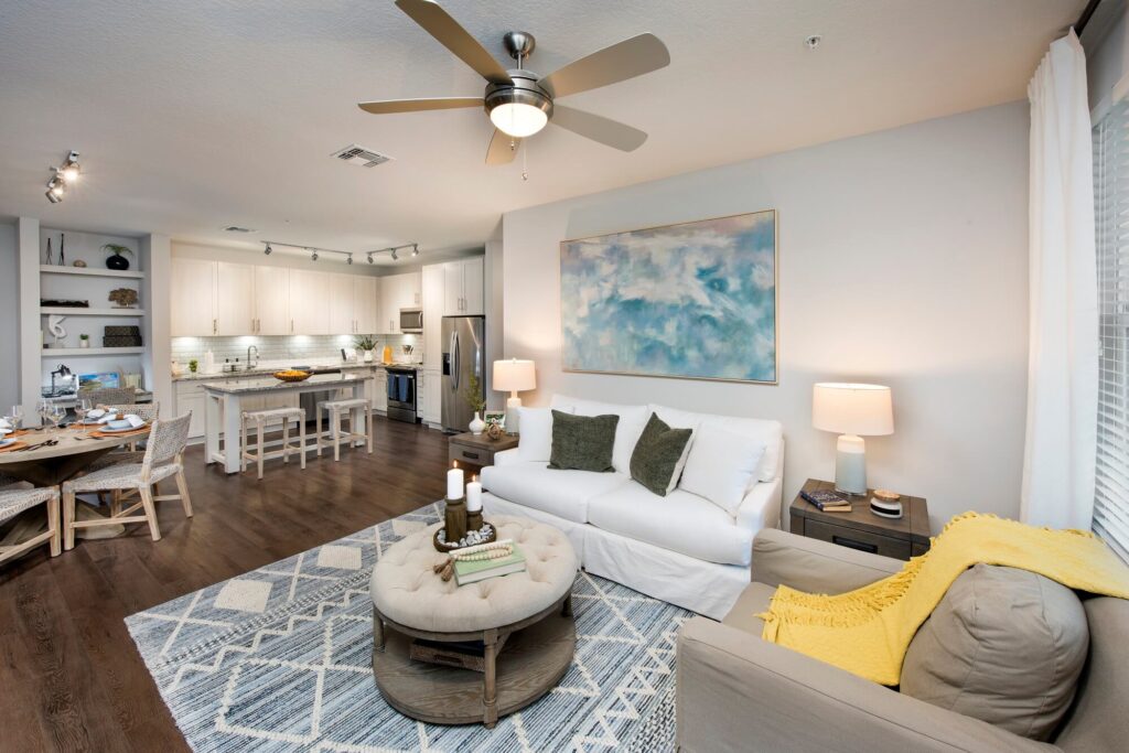 Modern, open-concept living room and kitchen with wood floors, neutral furniture, a white sofa, an armchair with a yellow throw, a patterned rug, round ottoman, wall art, and bright, natural light from large windows.