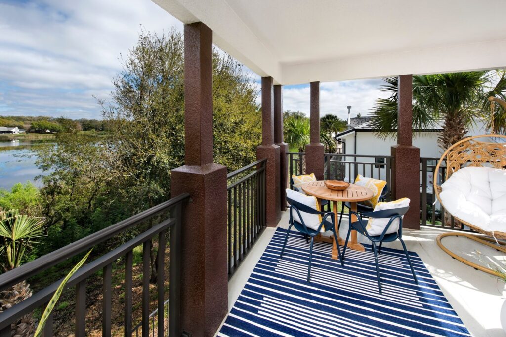 A covered balcony with brown columns features a round wooden table, four chairs with cushions, a hanging chair, and a striped blue and white rug. The balcony overlooks trees and a body of water under a partly cloudy sky.