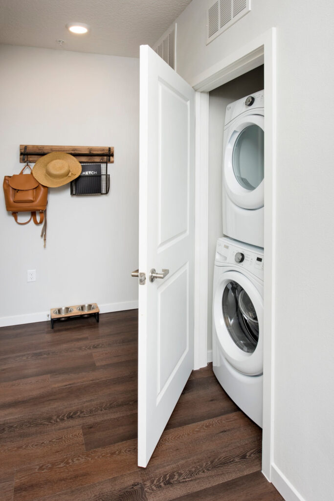 A white door is open to reveal a stacked washer and dryer in a closet. The room has dark wood floors, a wall-mounted shelf with hats and a bag, and a pet food and water bowl on the floor.