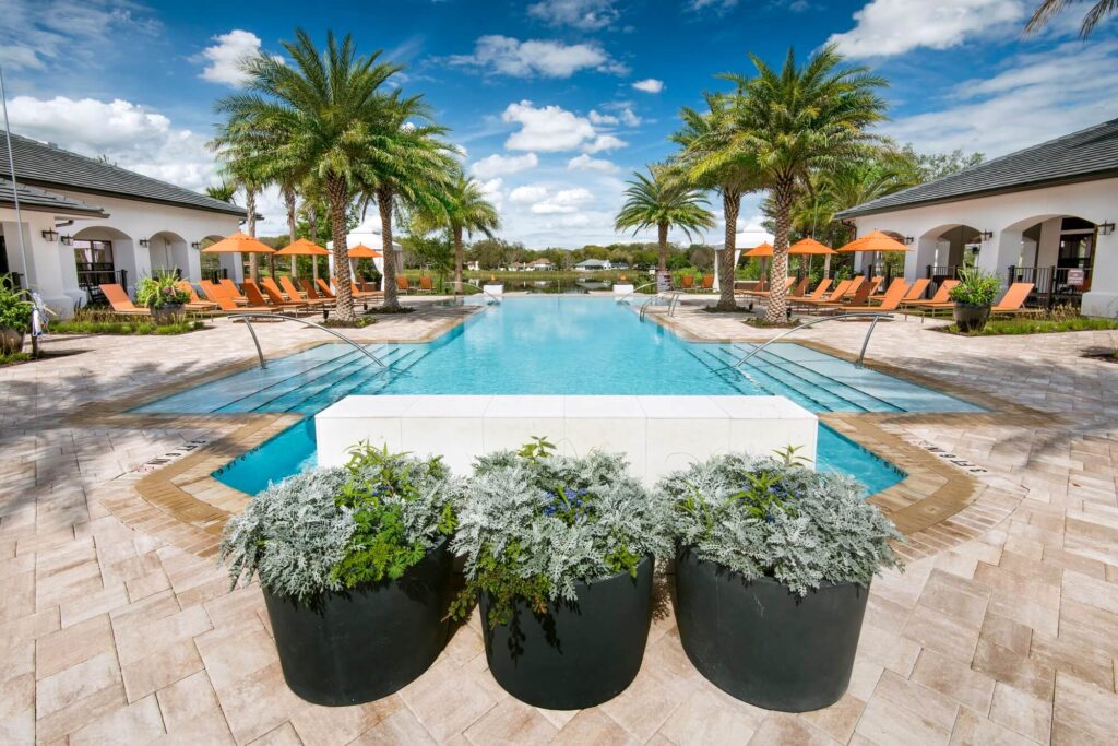 A luxurious outdoor pool flanked by palm trees, orange lounge chairs, and white buildings under a partly cloudy blue sky, with three large potted plants in the foreground.