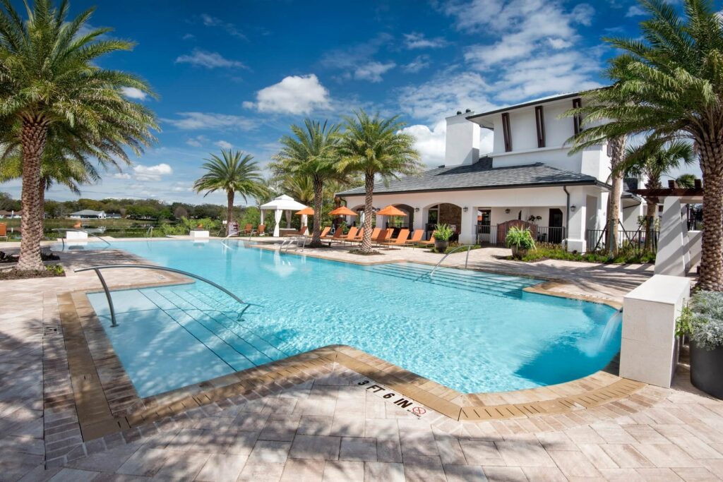 Outdoor swimming pool with clear blue water, surrounded by palm trees, lounge chairs, and umbrellas. A modern white clubhouse with large windows is in the background under a bright blue sky with scattered clouds.