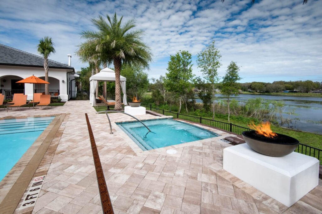 A resort-style patio features a pool, a hot tub, lounge chairs, a cabana, and two fire bowls. Palm trees and greenery surround the area, with a lake and blue sky with clouds in the background.
