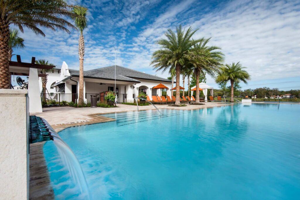 Large swimming pool with clear blue water, palm trees, orange lounge chairs, and a white building under a partly cloudy sky, creating a relaxing resort-like atmosphere.
