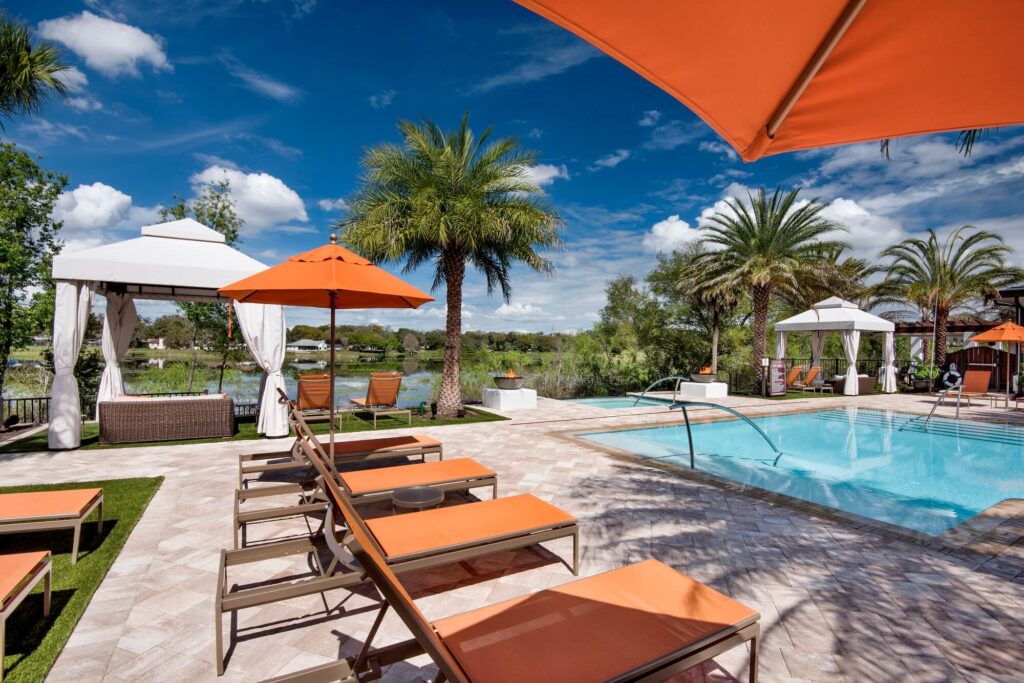 A sunny outdoor pool area with orange lounge chairs and umbrellas, palm trees, cabanas, and a view of a lake and blue sky with clouds in the background.